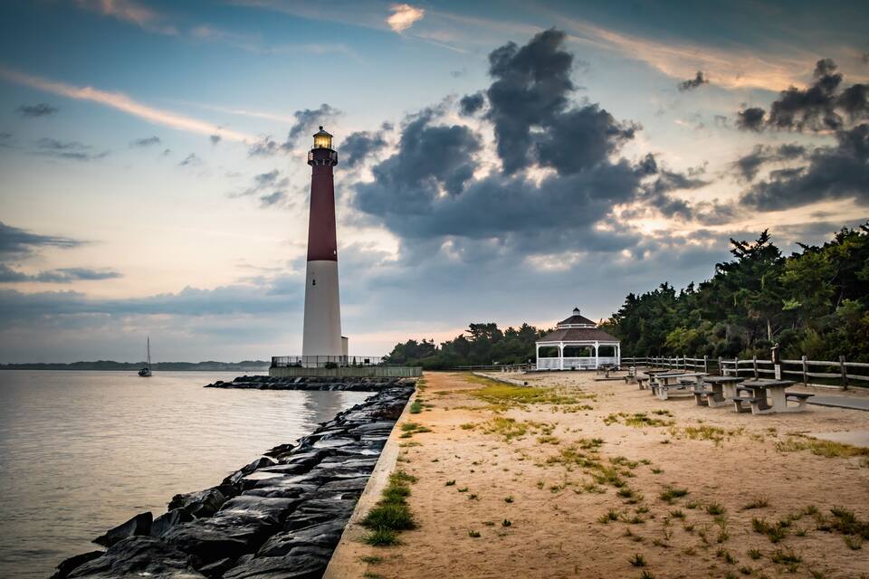 A lighthouse, a sandy beach, a wooden fence, and a gazebo by the water.