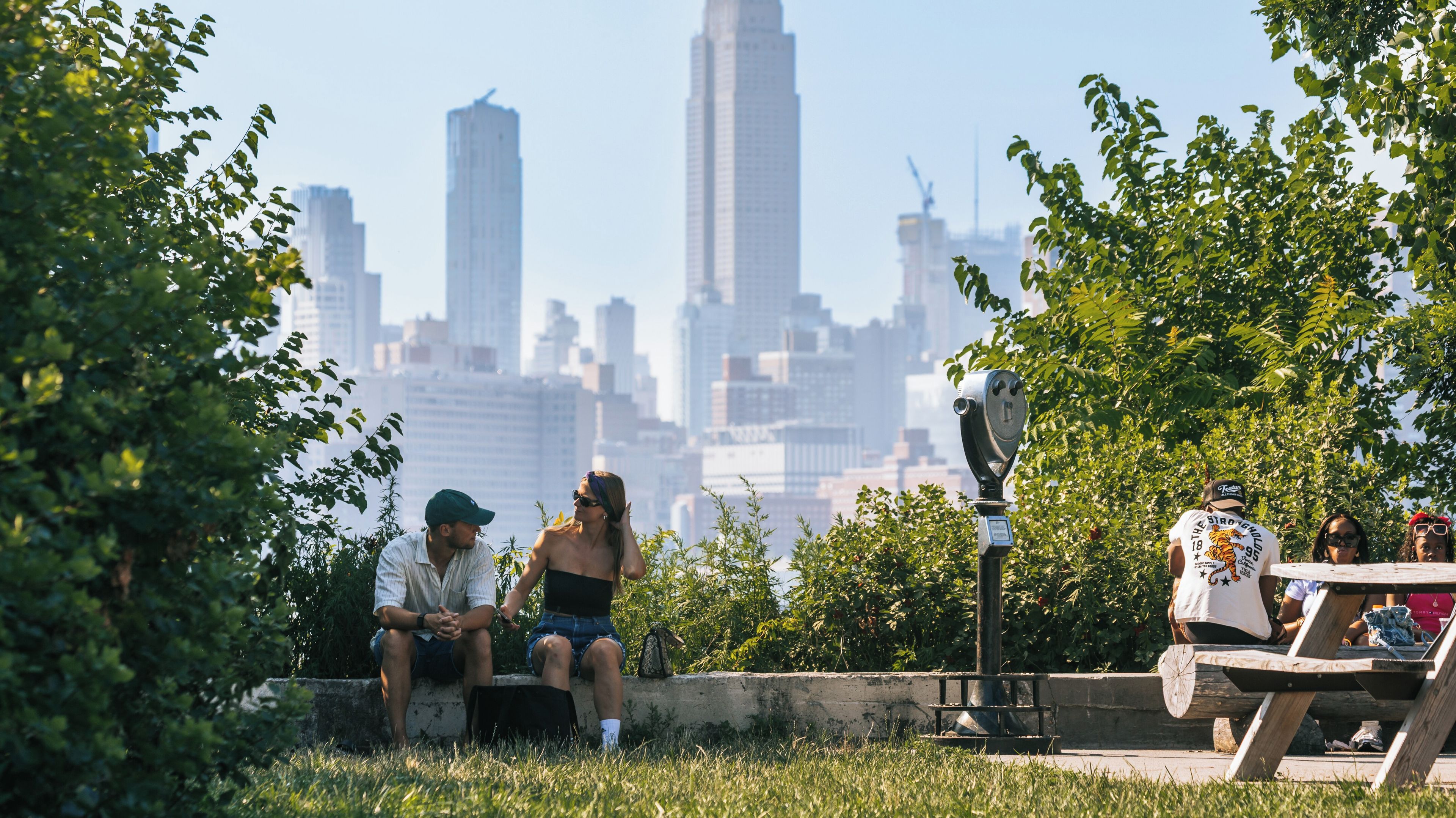 A park with people sitting on benches, a metal parking meter, and a city skyline in the background.
