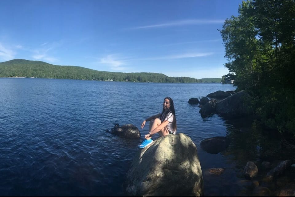 A person sitting on a rock by a lake with a forested hill in the background.