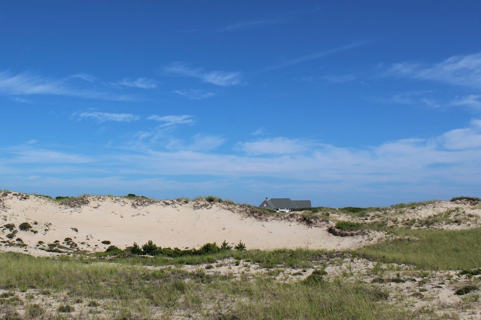 Un paysage de dunes de sable avec une végétation clairsemée et une maison qui se profile à l’horizon, sous un ciel bleu dégagé.