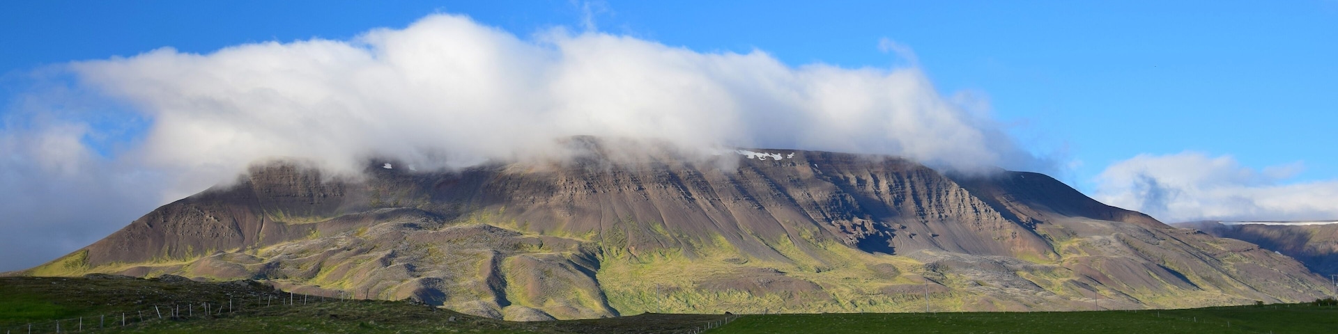 A mountain with a grassy foreground and a partly cloudy sky.