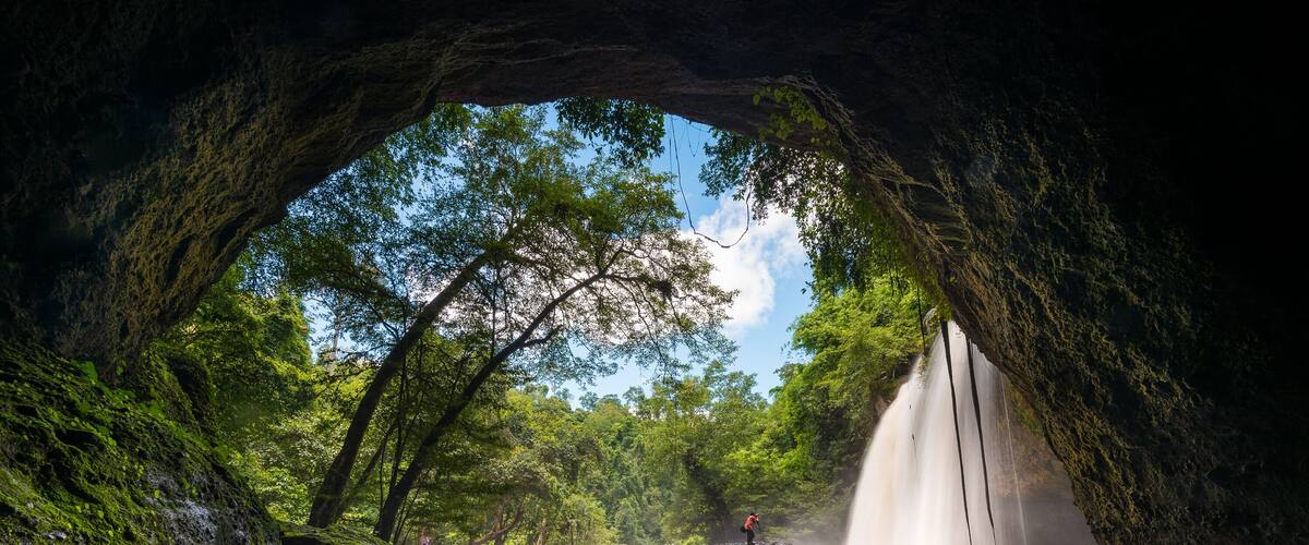 A cave entrance with a waterfall, surrounded by lush greenery and moss-covered rocks.