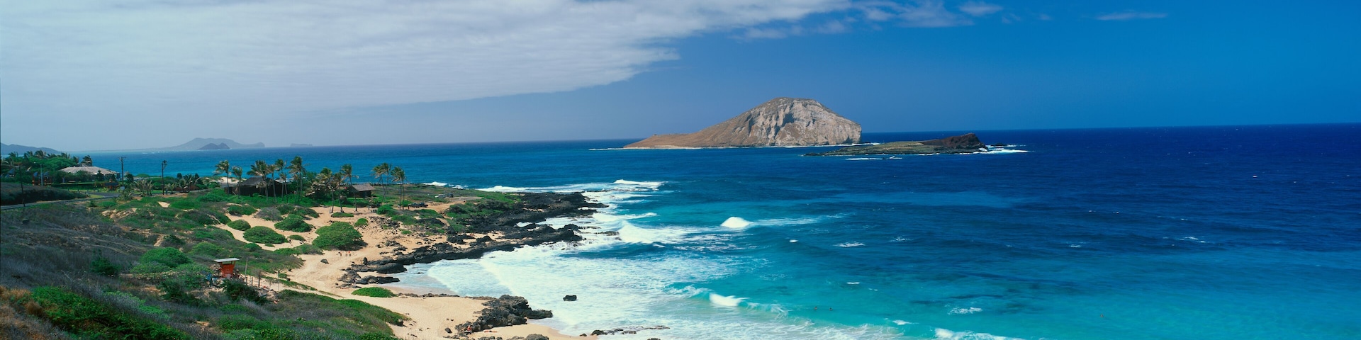Paisagem costeira com uma colina rochosa imponente, oceano azul cristalino, praia de areia e vegetação rasteira.