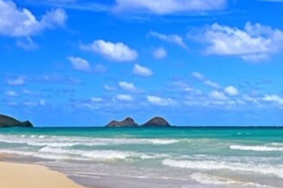 A beach with clear turquoise water, sandy shore, and distant rocky islands under a blue sky with scattered clouds.