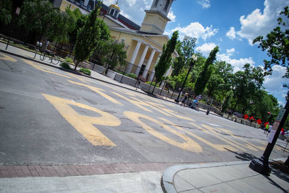 Uma praça urbana com uma grande placa de 'BUS STOP', um edifício histórico com colunas e um céu azul límpido.