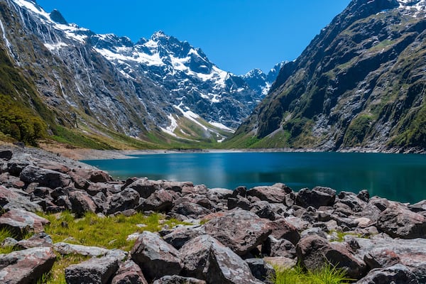 A mountainous landscape with a clear blue lake surrounded by rocky terrain and snow-capped peaks.