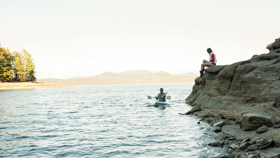 A kayaker on a calm lake with a rocky shore and a person sitting on a rock.