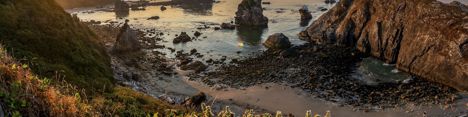 A coastal landscape with rocky formations, a beach, and a sunset.
