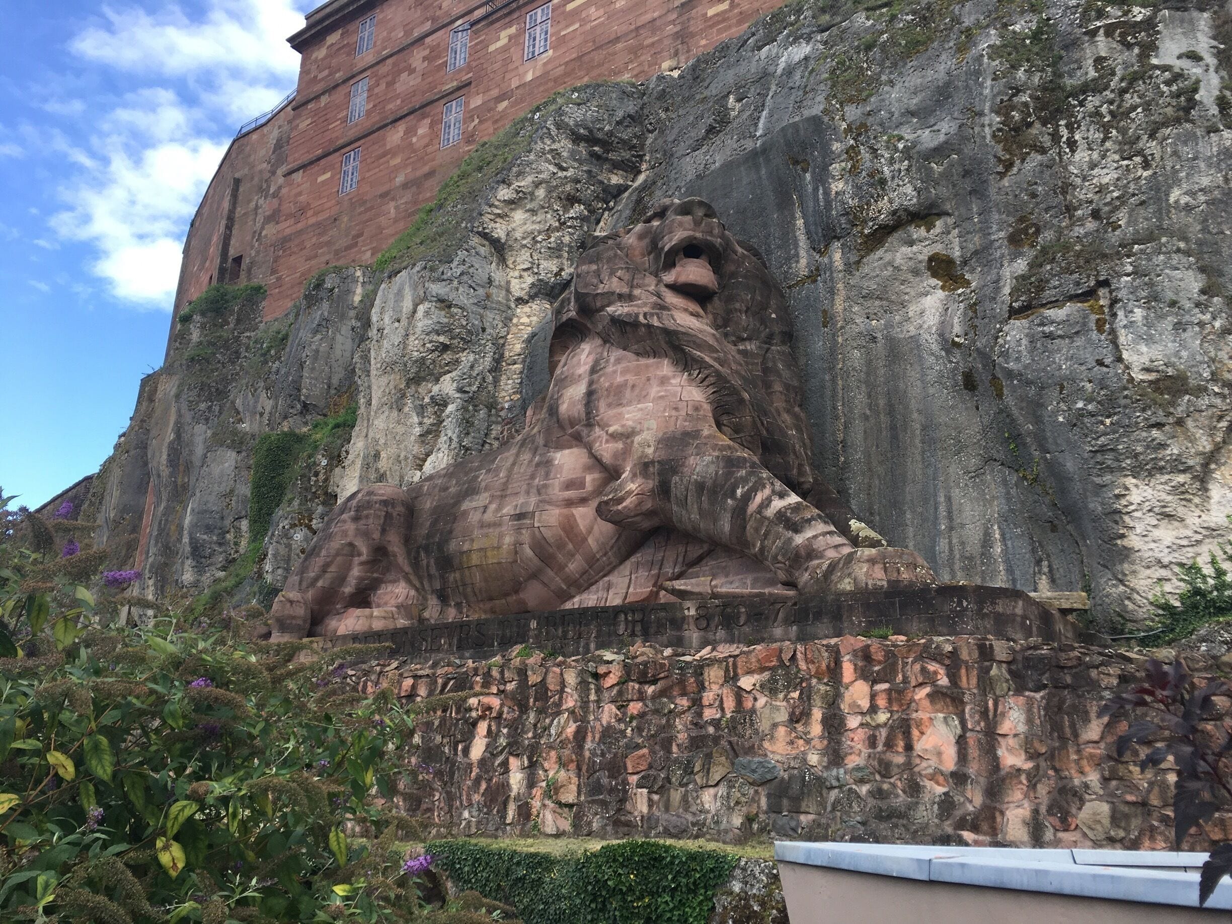 A large rock lion statue in a garden with a building in the background.