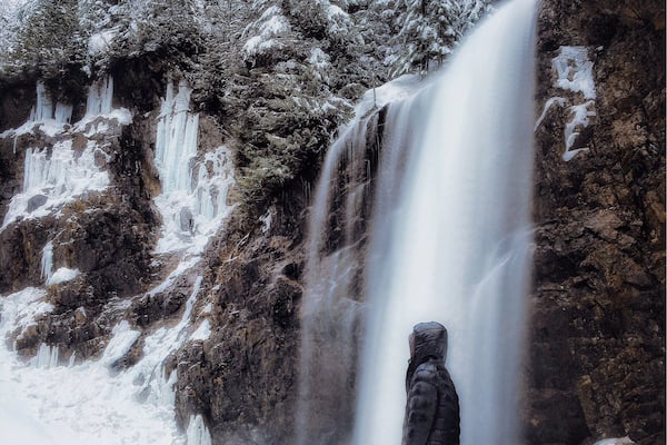 Une personne se tient devant une cascade, avec des arbres couverts de neige en arrière-plan.