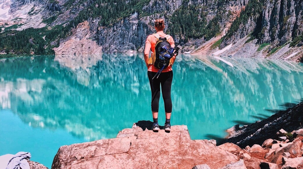 Una persona de pie sobre un promontorio rocoso con vistas a un lago turquesa y montañas al fondo.