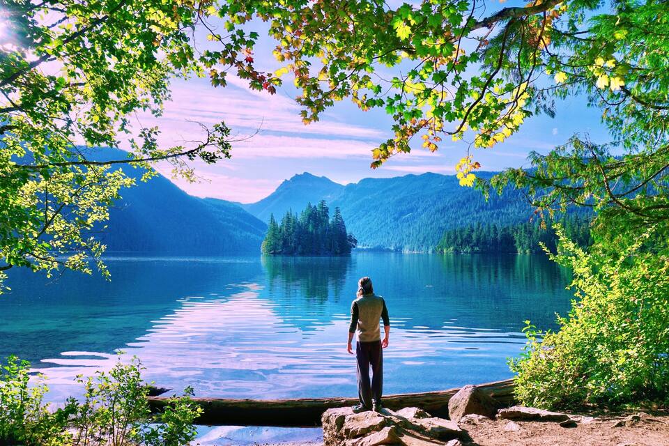 Una persona de pie junto a un lago tranquilo, rodeada de montañas y árboles.