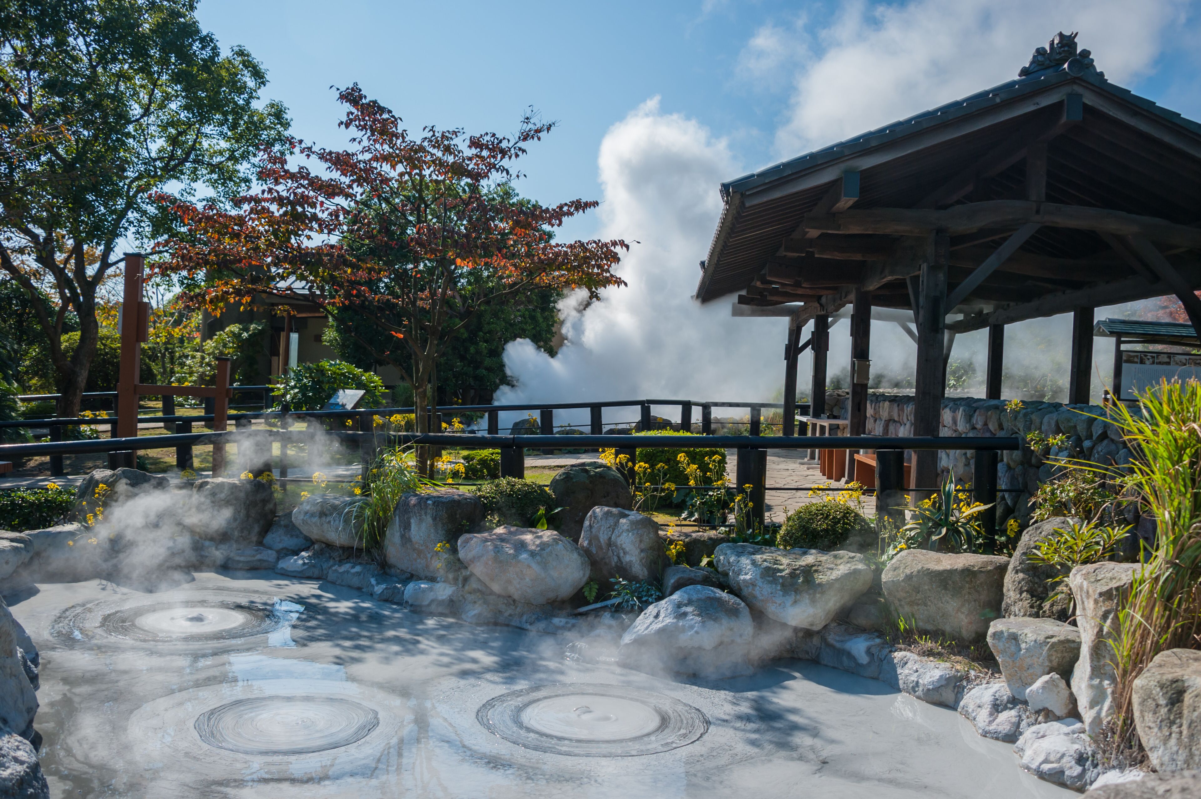 Uma área de águas termais com vapor subindo, rodeada por pedras e árvores, e um pavilhão de madeira tradicional.