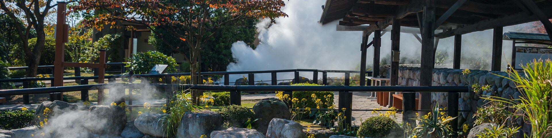 Uma área de águas termais com vapor subindo, rodeada por pedras e árvores, e um pavilhão de madeira tradicional.