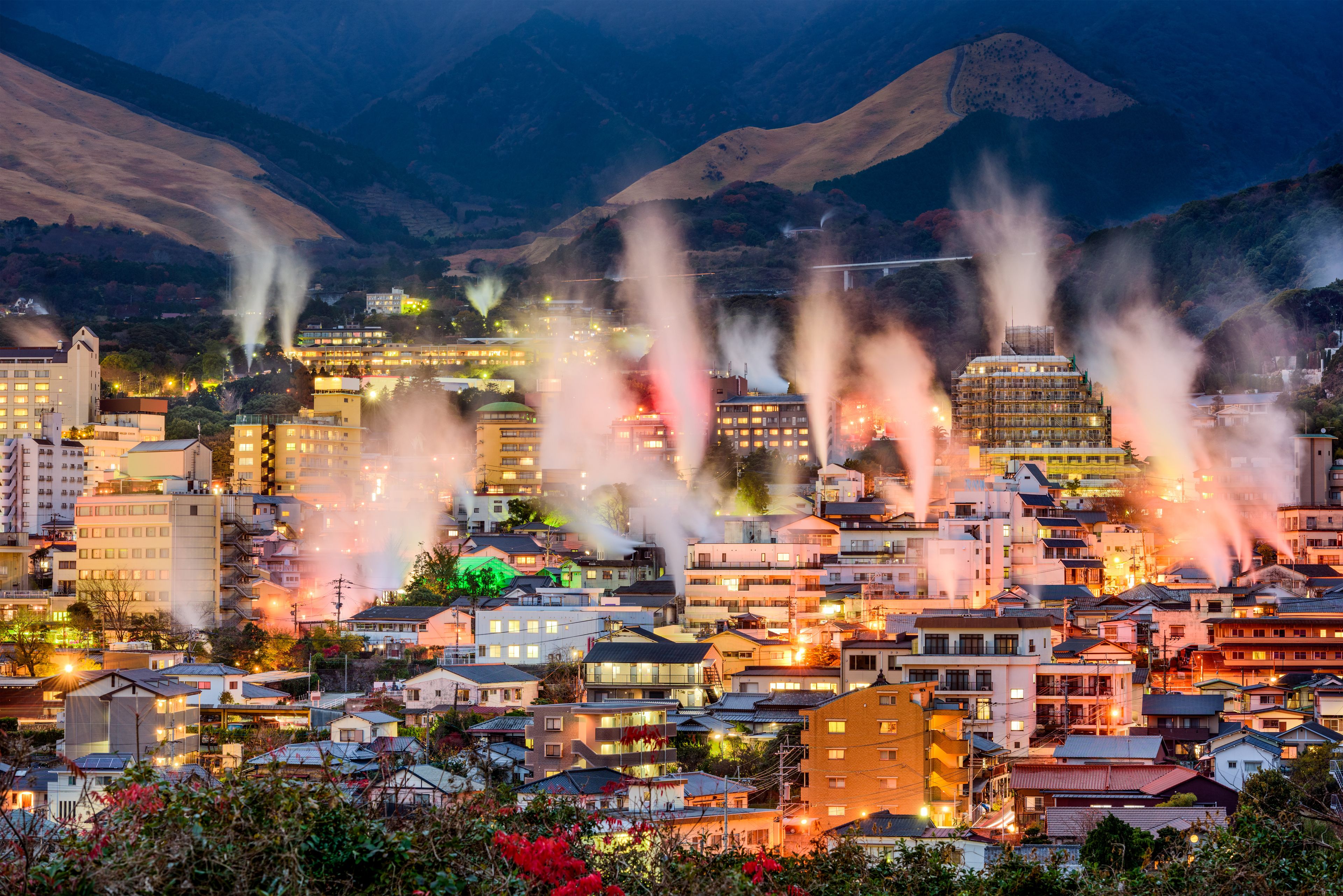 A cityscape at dusk with buildings, smoke plumes, and a mountainous backdrop.