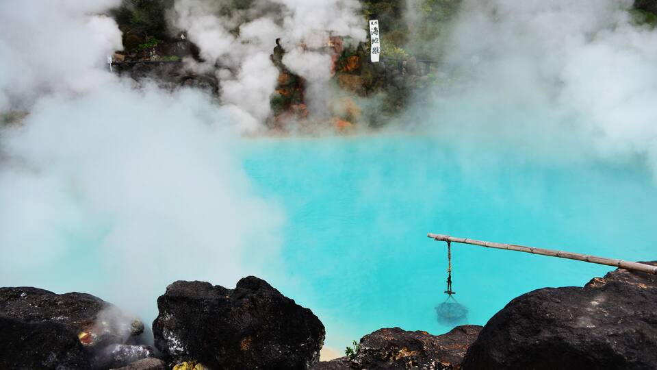 A geothermal hot spring with clear turquoise water, surrounded by dark volcanic rocks and emitting steam.