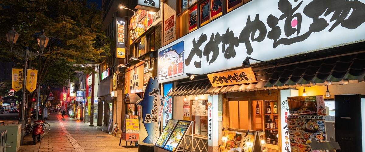 A nighttime street with illuminated signs and storefronts, including a sushi restaurant.