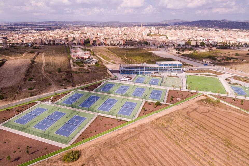 An aerial view of a sports complex with multiple tennis courts, surrounded by fields and a cityscape in the background.