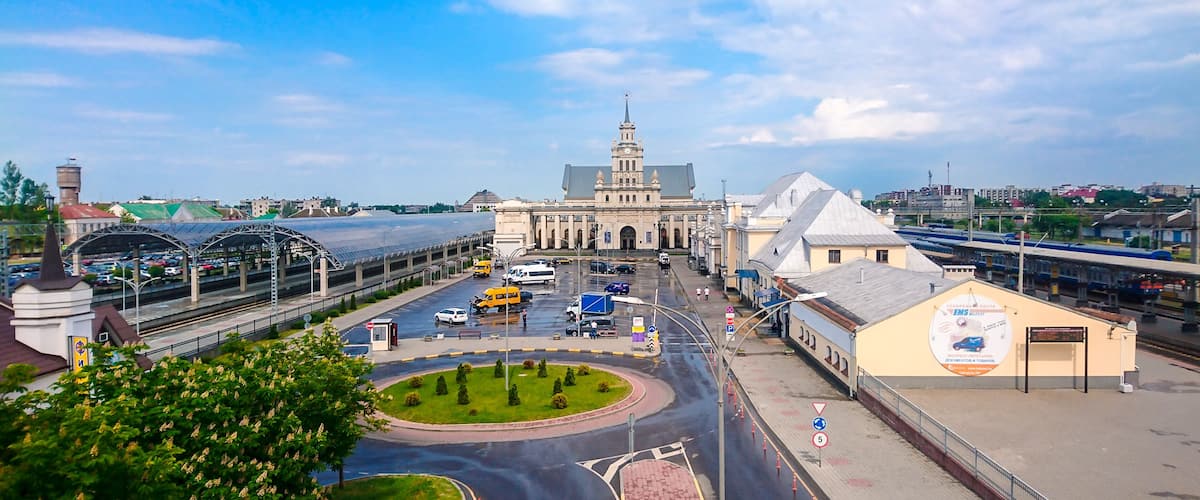 A cityscape with a prominent building with a clock tower, a train station, and a roundabout with greenery.