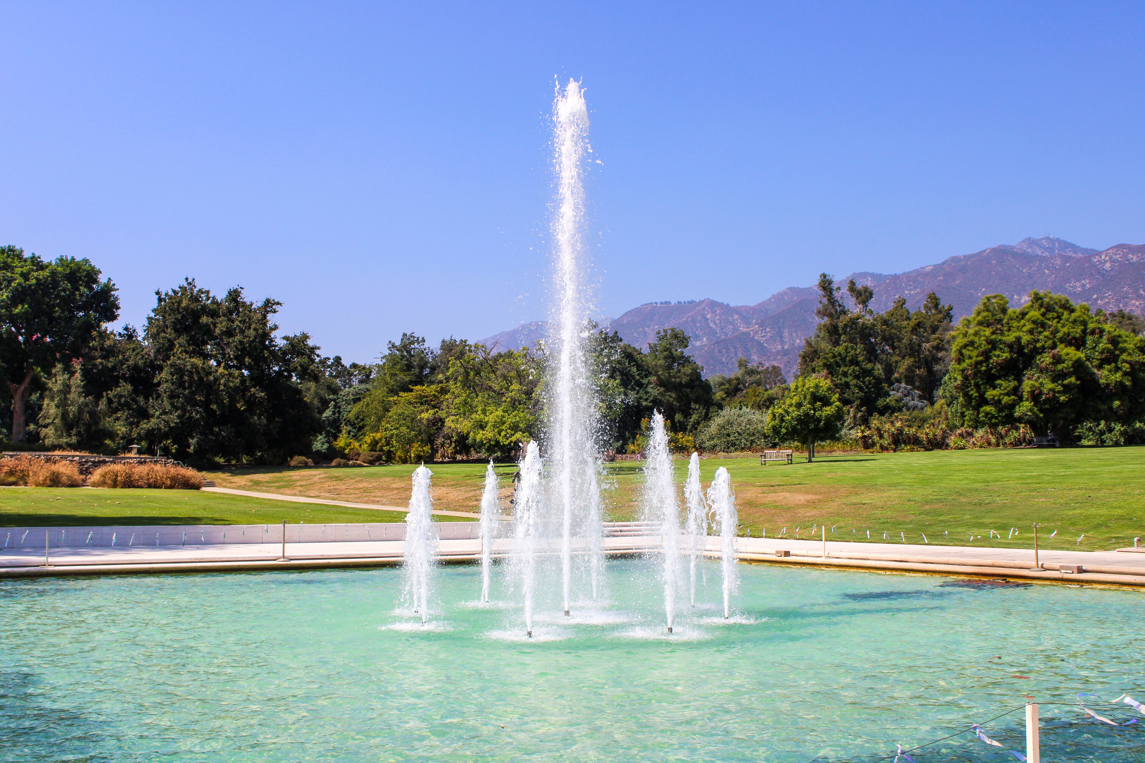 a gorgeous summer landscape at the LA Arboretum with a water fountain, lush green trees, plants and grass, mountains and blue sky in Arcadia California USA