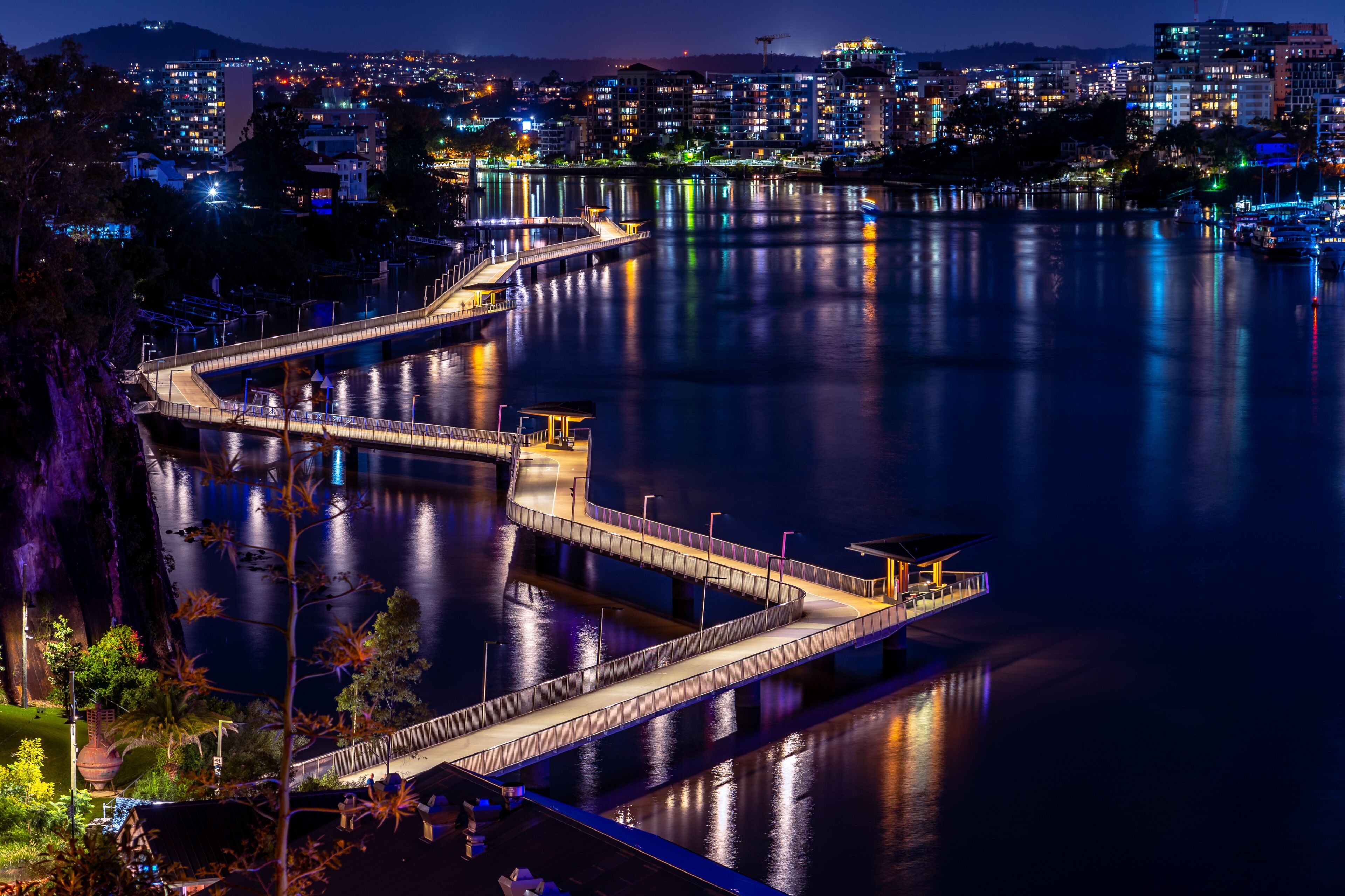 Un paisaje urbano nocturno con un puente iluminado sobre aguas tranquilas, que reflejan las luces de la ciudad.