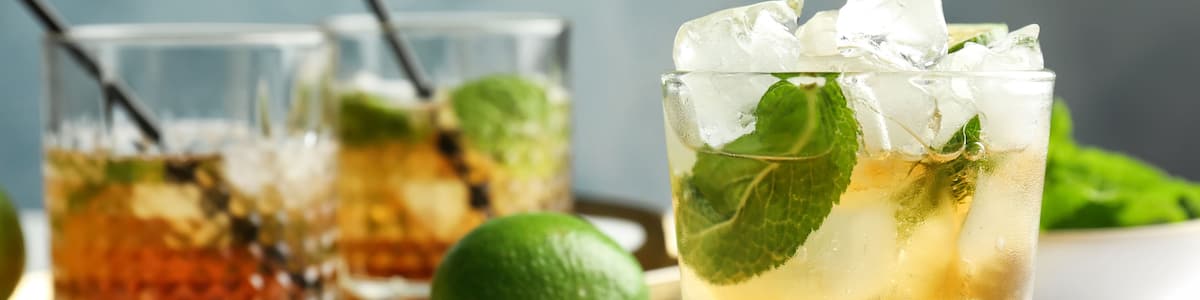 Zwei Gläser mit hellem Getränk, Eiswürfeln, Minzblättern, einer Limette und einem Löffel auf weißer Oberfläche.