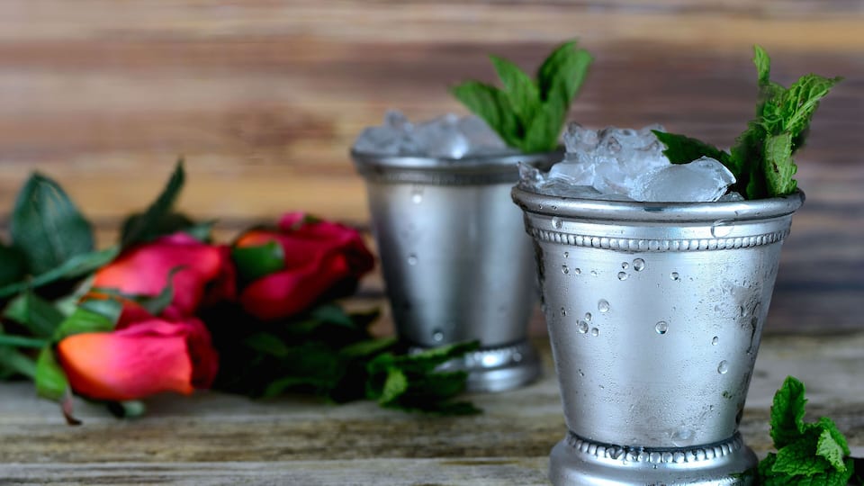 Two metallic cups with ice and mint leaves, accompanied by red roses and green leaves on a wooden surface.