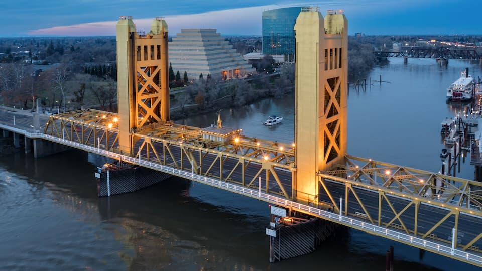 A steel bridge with two towers over a river, a boat on the water, and buildings in the background.