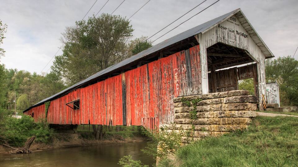 A red covered bridge with the inscription 'Rosette Bridge 1910' and a stone wall beside a calm river.