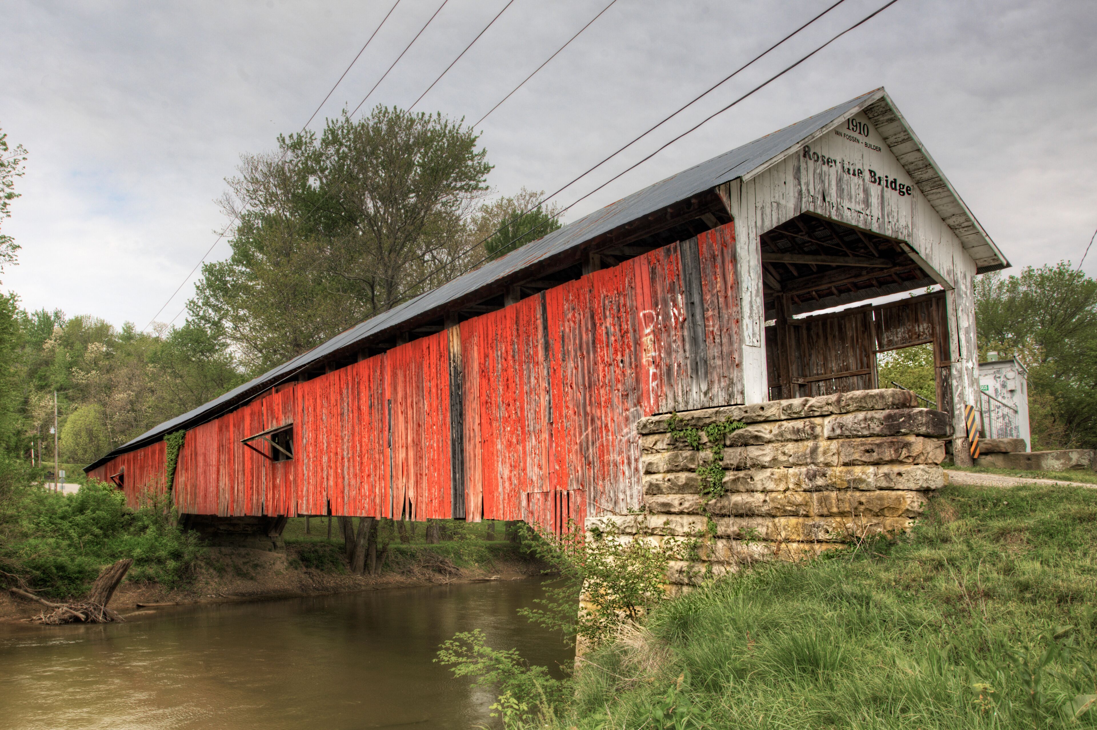 A red covered bridge with the inscription 'Rosette Bridge 1910' and a stone wall beside a calm river.