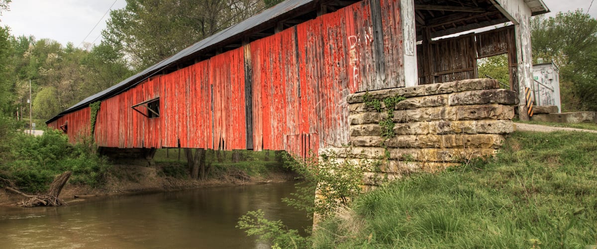 A red covered bridge with the inscription 'Rosette Bridge 1910' and a stone wall beside a calm river.