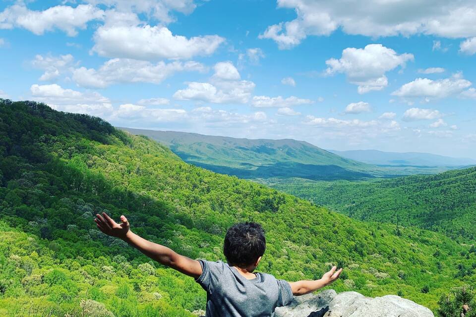 Una persona sentada sobre una roca con los brazos extendidos, contemplando un valle verde exuberante y colinas distantes bajo un cielo parcialmente nublado.