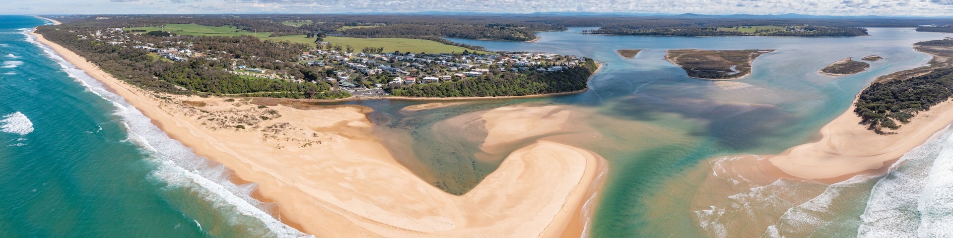 An aerial view of a coastal town with a beach, clear waters, and surrounding greenery.