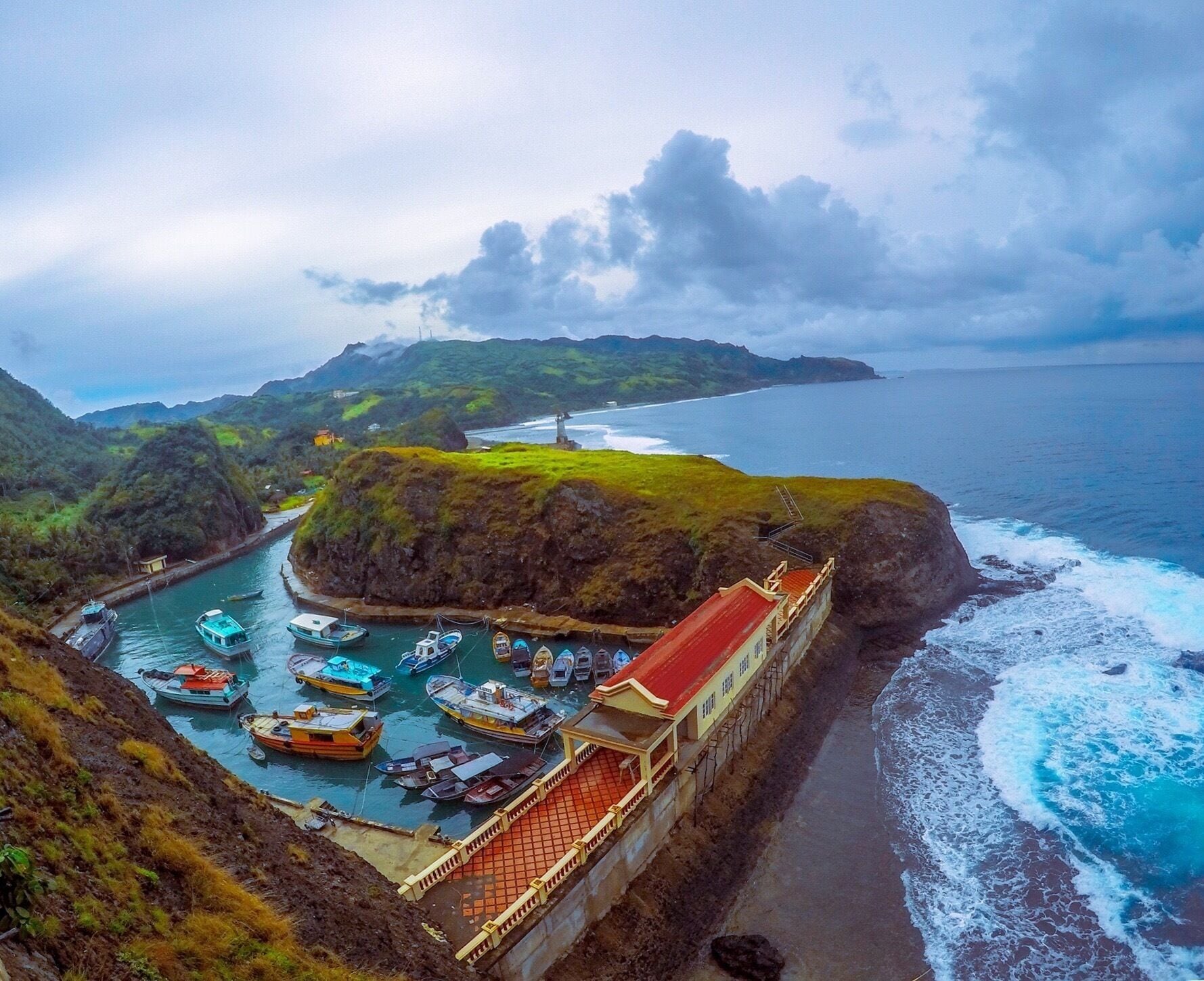 A coastal village with a red-roofed building, a harbor with small boats, and a lighthouse in the distance.