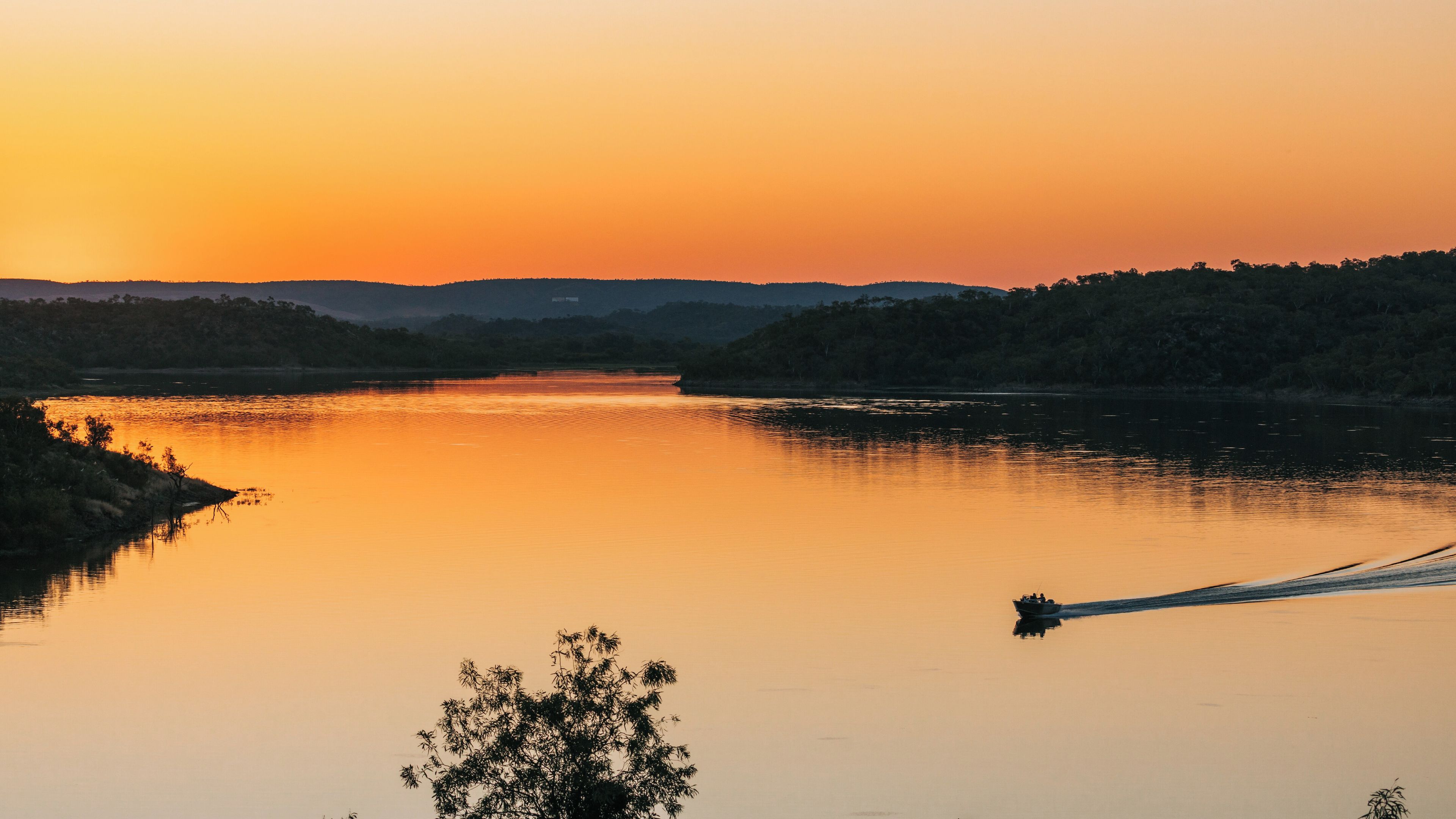 A calm river at sunset with a boat creating a wake, surrounded by hills and trees.