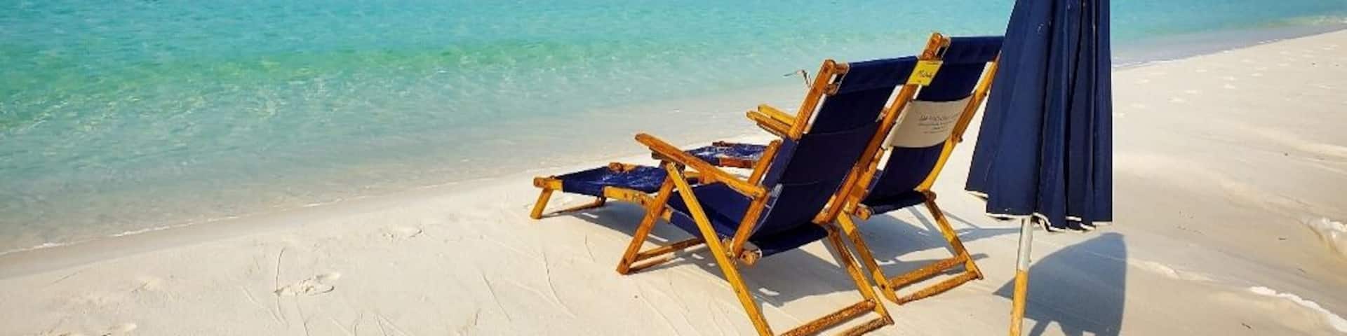 A beach with two wooden lounge chairs and a dark blue umbrella on the sand.