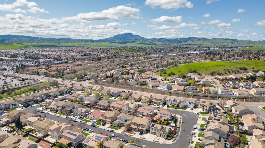 Aerial view of a suburban neighborhood and shopping center in Antioch, California, on a clear spring day with scenic hills, green fields, and residential homes with solar panels.