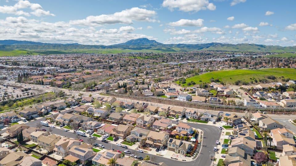 Aerial view of a suburban neighborhood and shopping center in Antioch, California, on a clear spring day with scenic hills, green fields, and residential homes with solar panels.