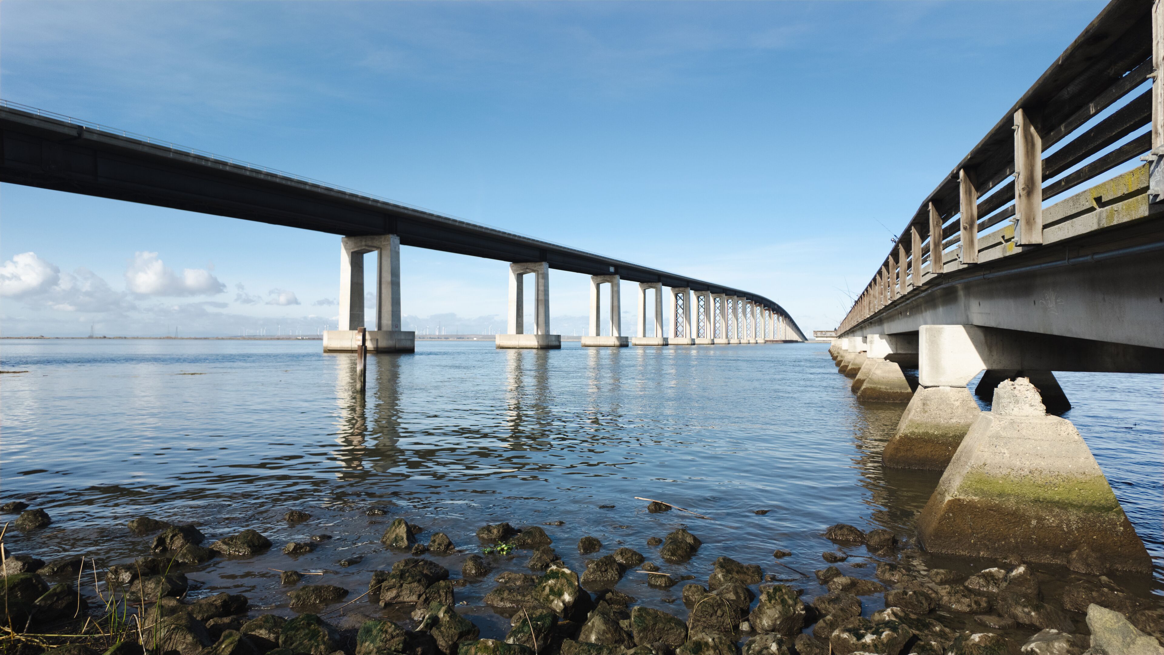 A bridge over calm water with a rocky shoreline and distant mountains.