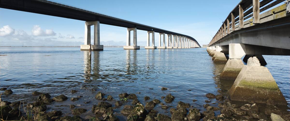 A bridge over calm water with a rocky shoreline and distant mountains.