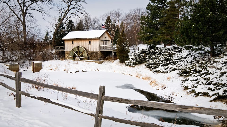 A wooden watermill with a large wheel, surrounded by snow-covered ground and trees.