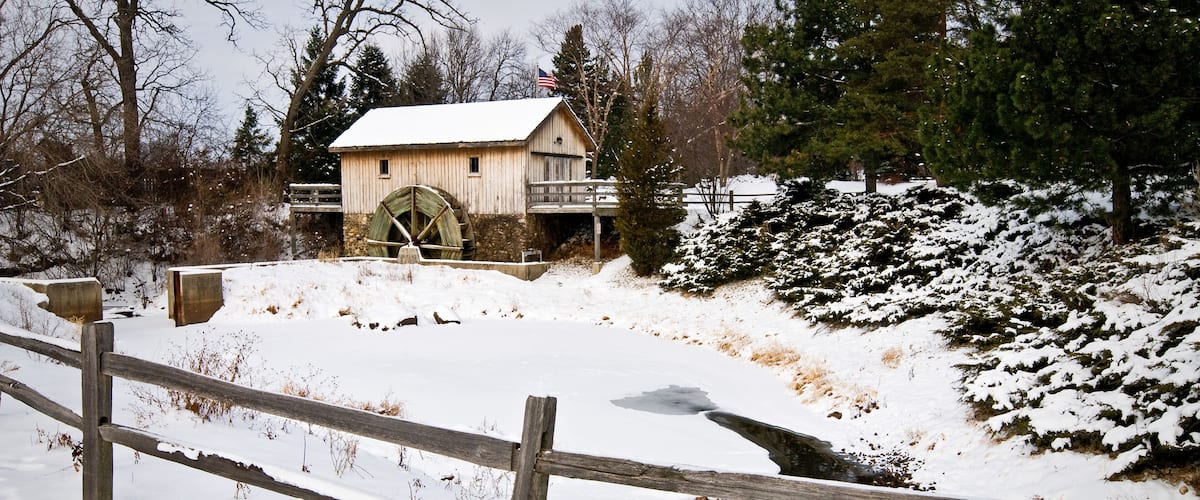 A wooden watermill with a large wheel, surrounded by snow-covered ground and trees.