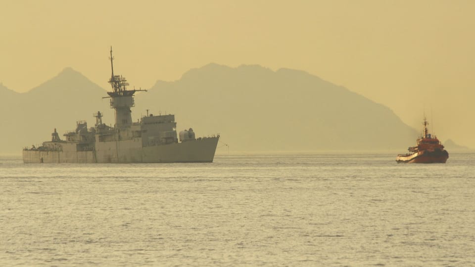 A large naval ship and a smaller tugboat in the ocean with mountains in the background.
