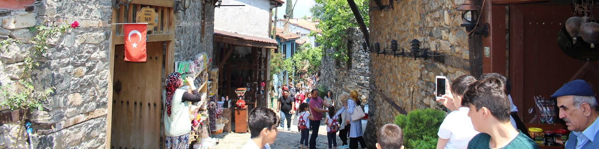 A narrow street with stone buildings, a shop with a Turkish flag, and people walking.