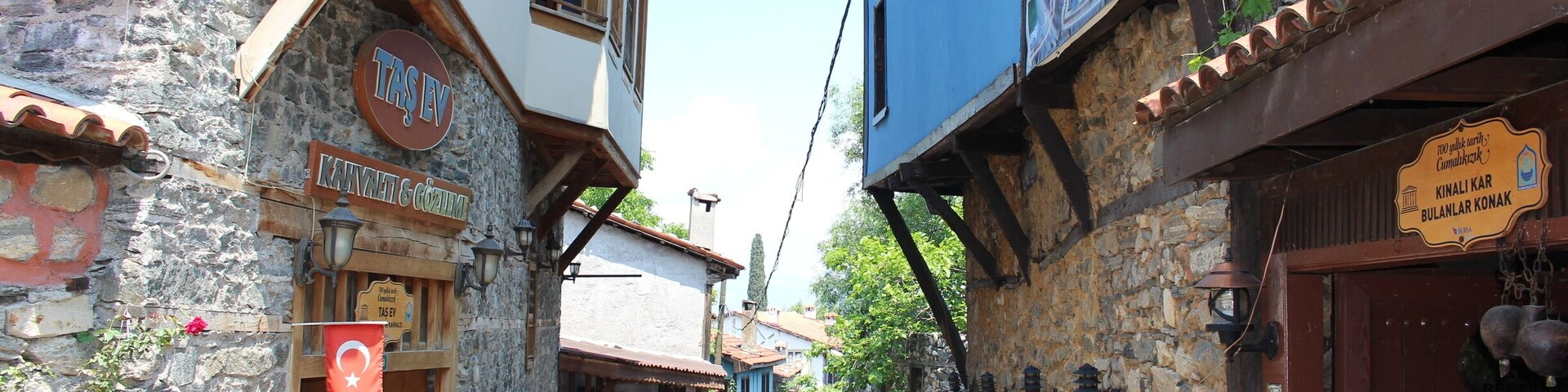 Uma rua estreita com edifícios de pedra, uma loja com a bandeira turca e pessoas caminhando.