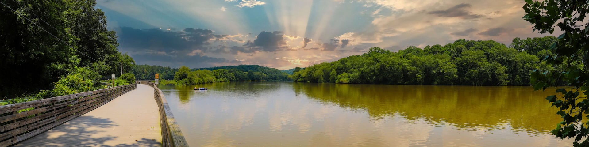 a panoramic shot of the still silky brown water of the Chattahoochee river with a long wooden boardwalk along the river and lush green trees with powerful clouds at Roswell Riverwalk in Georgia