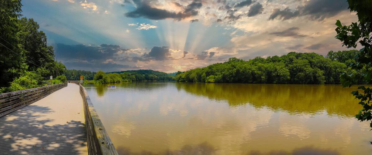 A calm river with a wooden bridge, surrounded by dense greenery and a cloudy sky.