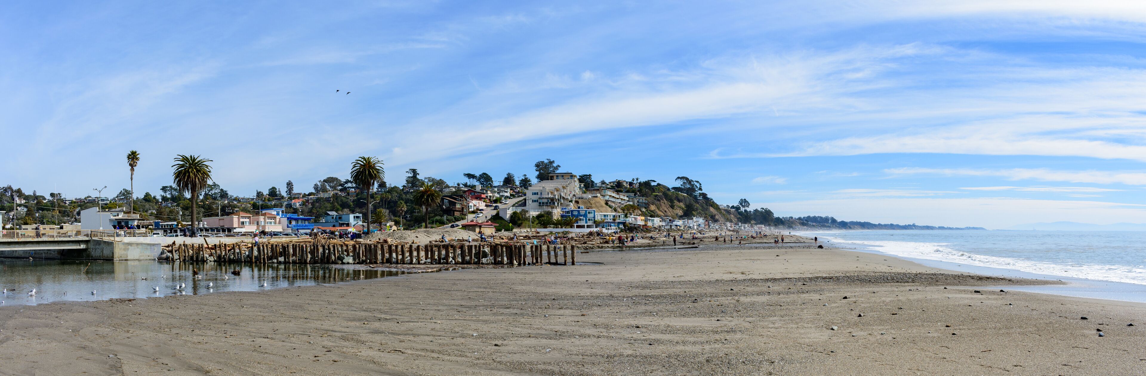 Scenic panoramic view of Rio Del Mar Beach near Santa Cruz California with a wooden pier stretching into water, flanked by coastal buildings and palms