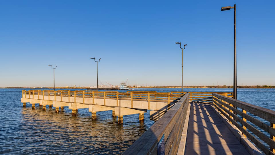 A wooden pier extending into calm waters, with a clear sky above and a distant industrial area in the background.