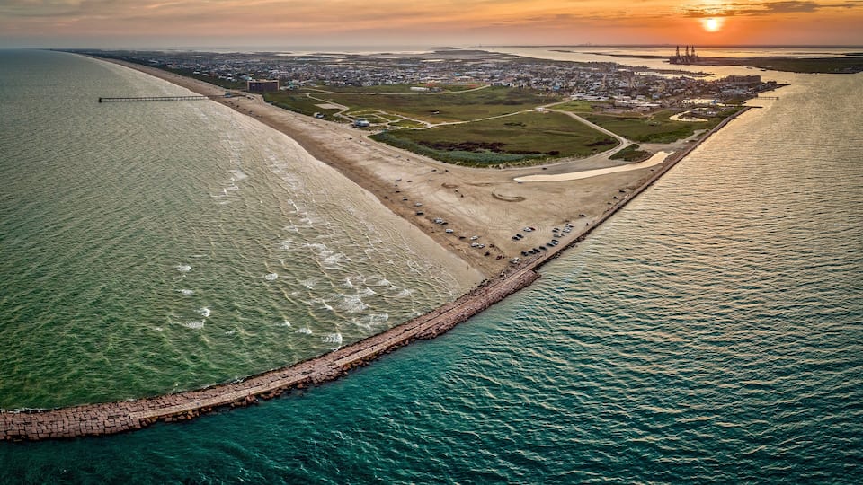 An aerial view of a coastal city with a long pier extending into the sea, a beach, and a large industrial complex in the distance.
